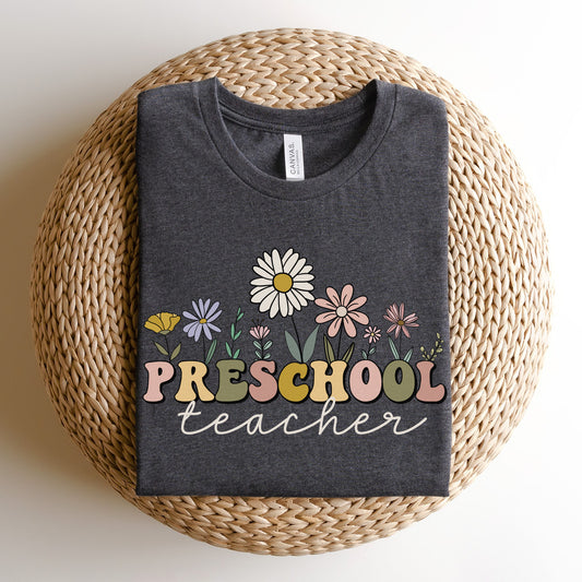 Tshirt that says "Preschool Teacher" Preschool is in a multicolored font and underneath it is 'teacher' in a light cursive text. Different colored and shaped wildflowers are coming out of the top of the word 'preschool'. Colored tshirt: dark grey heather 