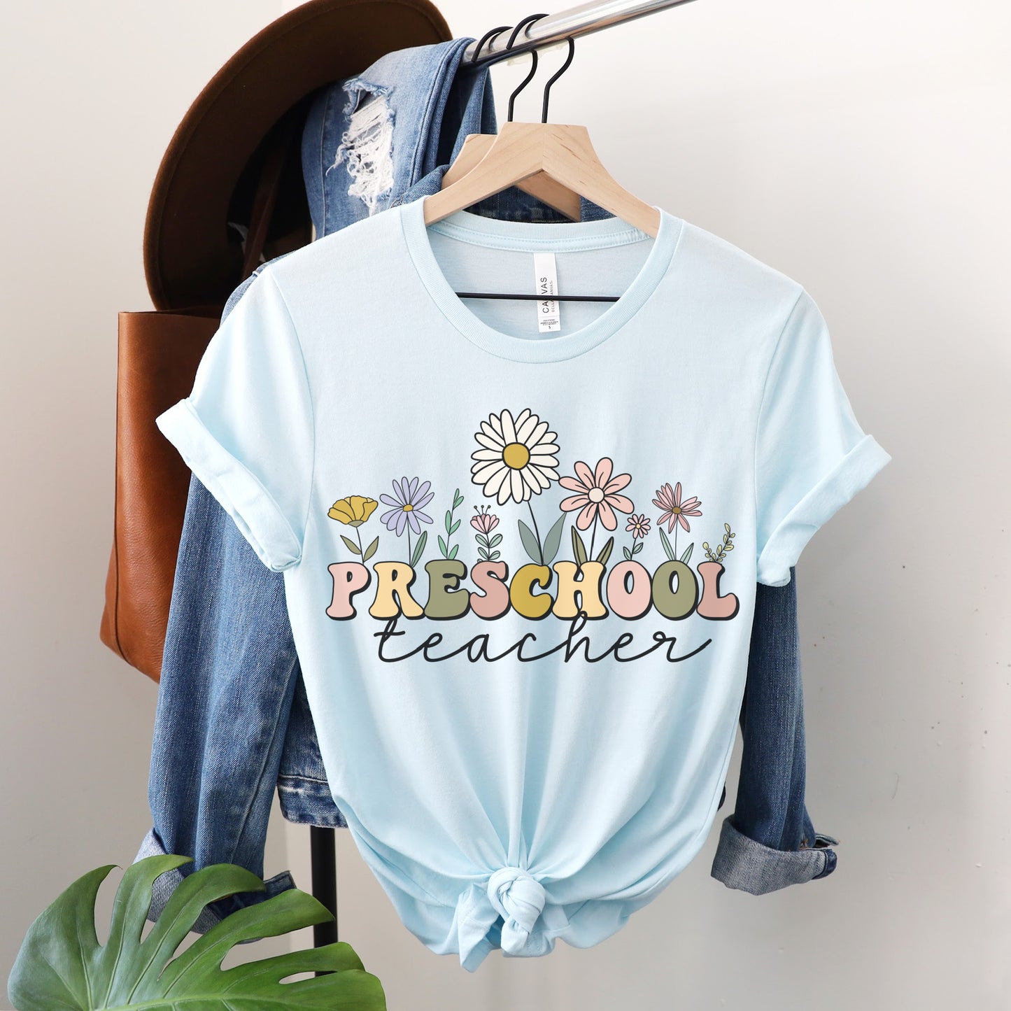 Tshirt that says "Preschool Teacher" Preschool is in a multicolored font and underneath it is 'teacher' in a black cursive text. Different colored and shaped wildflowers are coming out of the top of the word 'preschool'. Colored tshirt: heather ice blue