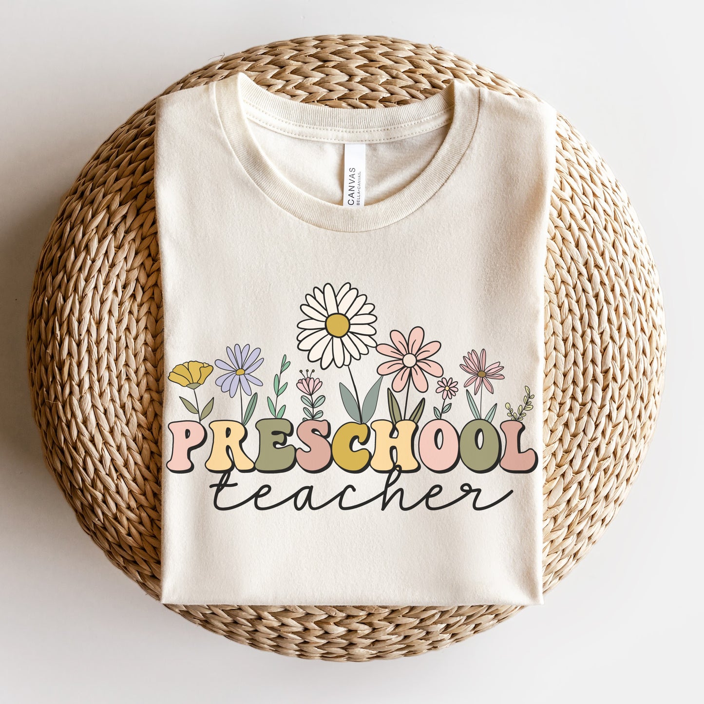 Tshirt that says "Preschool Teacher" Preschool is in a multicolored font and underneath it is 'teacher' in a black cursive text. Different colored and shaped wildflowers are coming out of the top of the word 'preschool'. Colored tshirt: natural