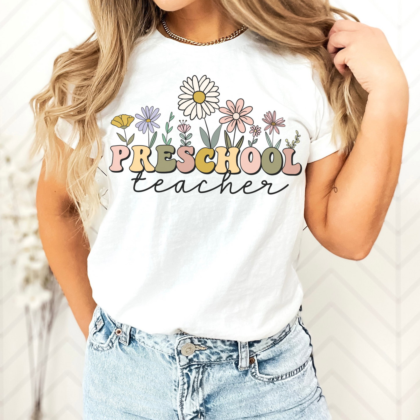 Tshirt that says "Preschool Teacher" Preschool is in a multicolored font and underneath it is 'teacher' in a black cursive text. Different colored and shaped wildflowers are coming out of the top of the word 'preschool'. Colored tshirt: white