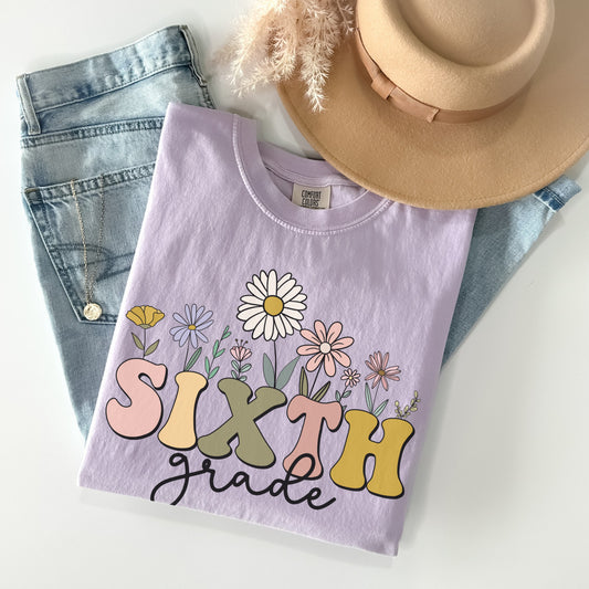 Tshirt that says "Sixth Grade". 'SIXTH' is in a bold, multicolored font with wildflowers coming out of the top of it. 'grade' is underneath in a black cursive font. Tshirt color: Orchid