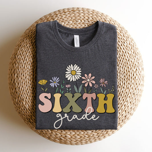 Tshirt that says "Sixth Grade" Sixth is in a multicolored font and underneath it is 'grade' in a light cursive font. Different colored and shaped wildflowers are coming out of the top of 'sixth'. Colored tshirt: dark grey heather 